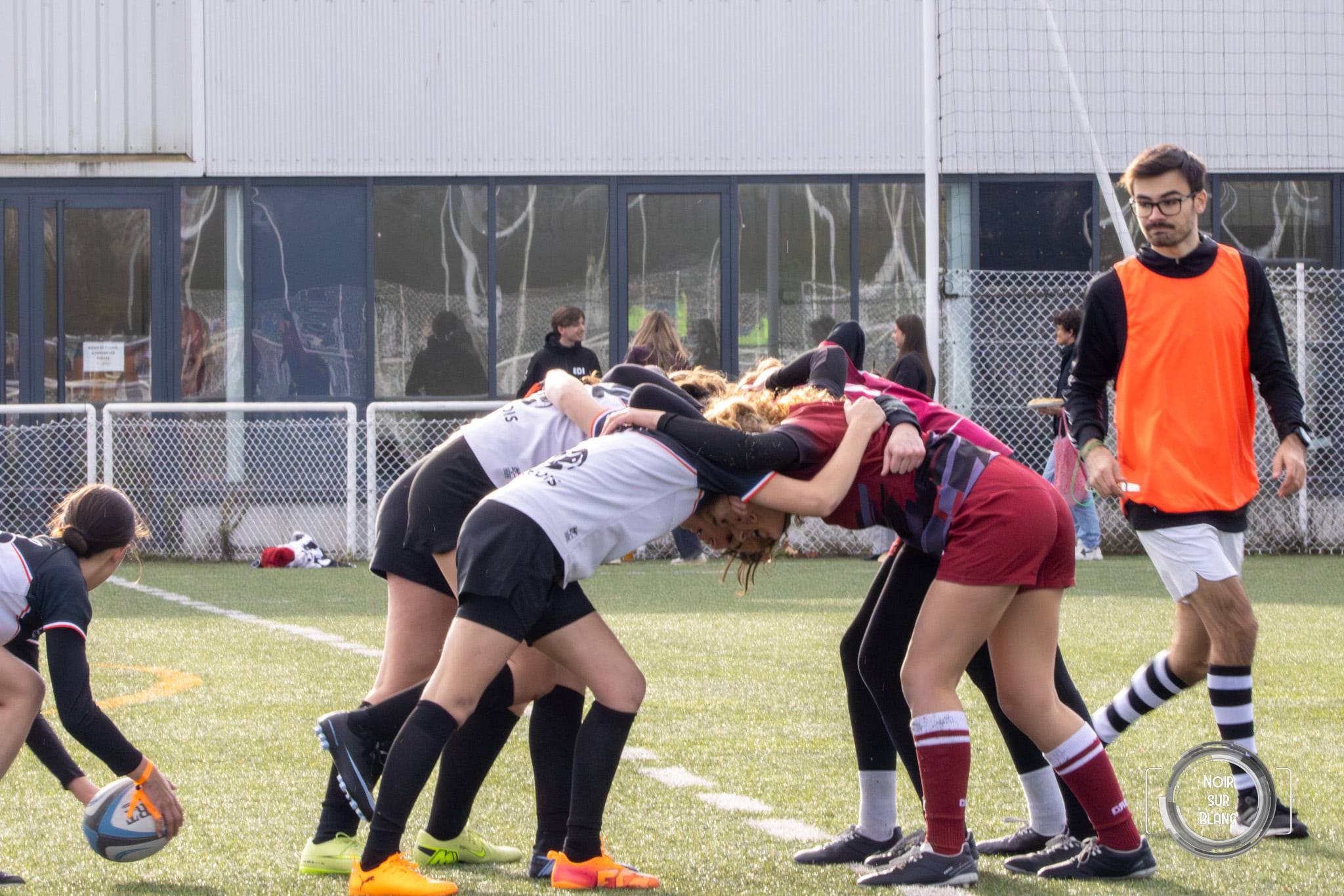 Sur un terrain en herbe, deux paquets de joueuses de rugby s'engagent dans une mêlée, l'arbitre en chasuble orange à côté d'elles.