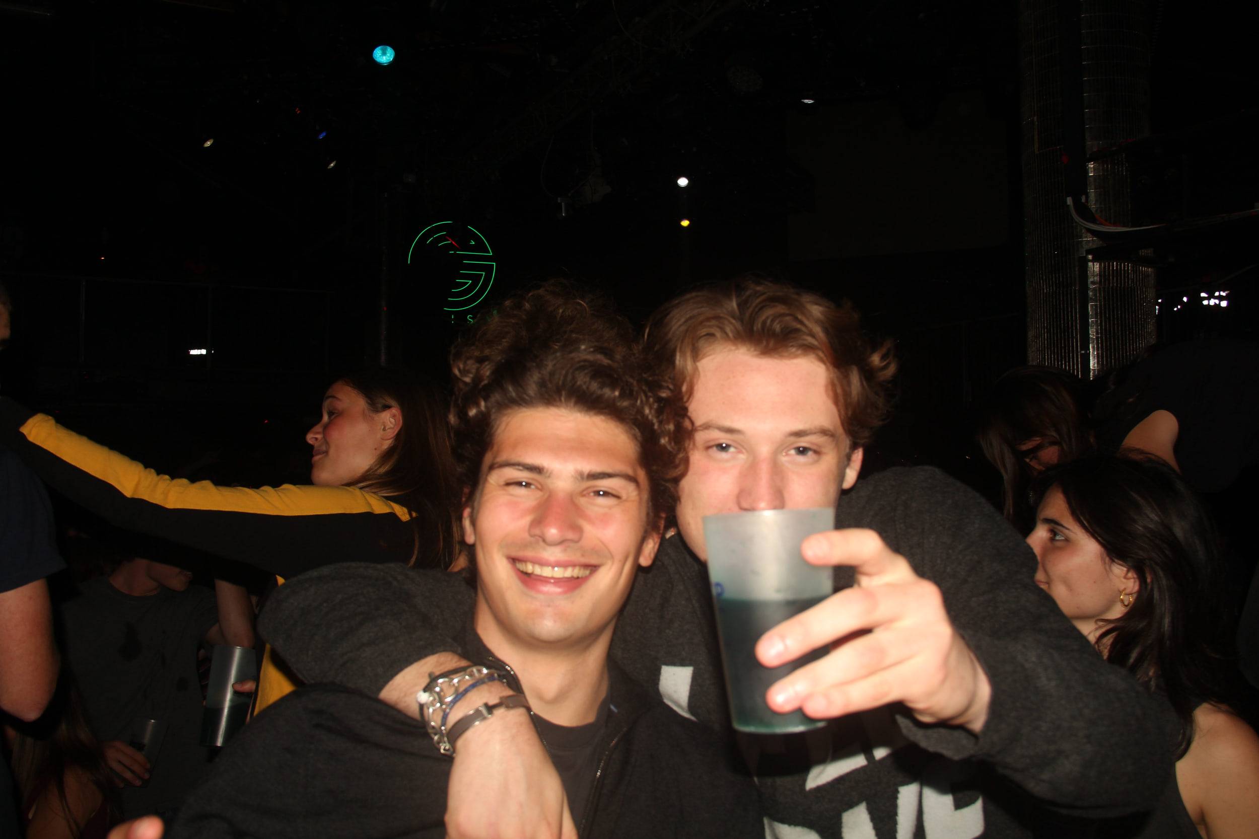 Deux amis posent bras dessus bras dessous, l'un tend son gobelet vers l'objectif.