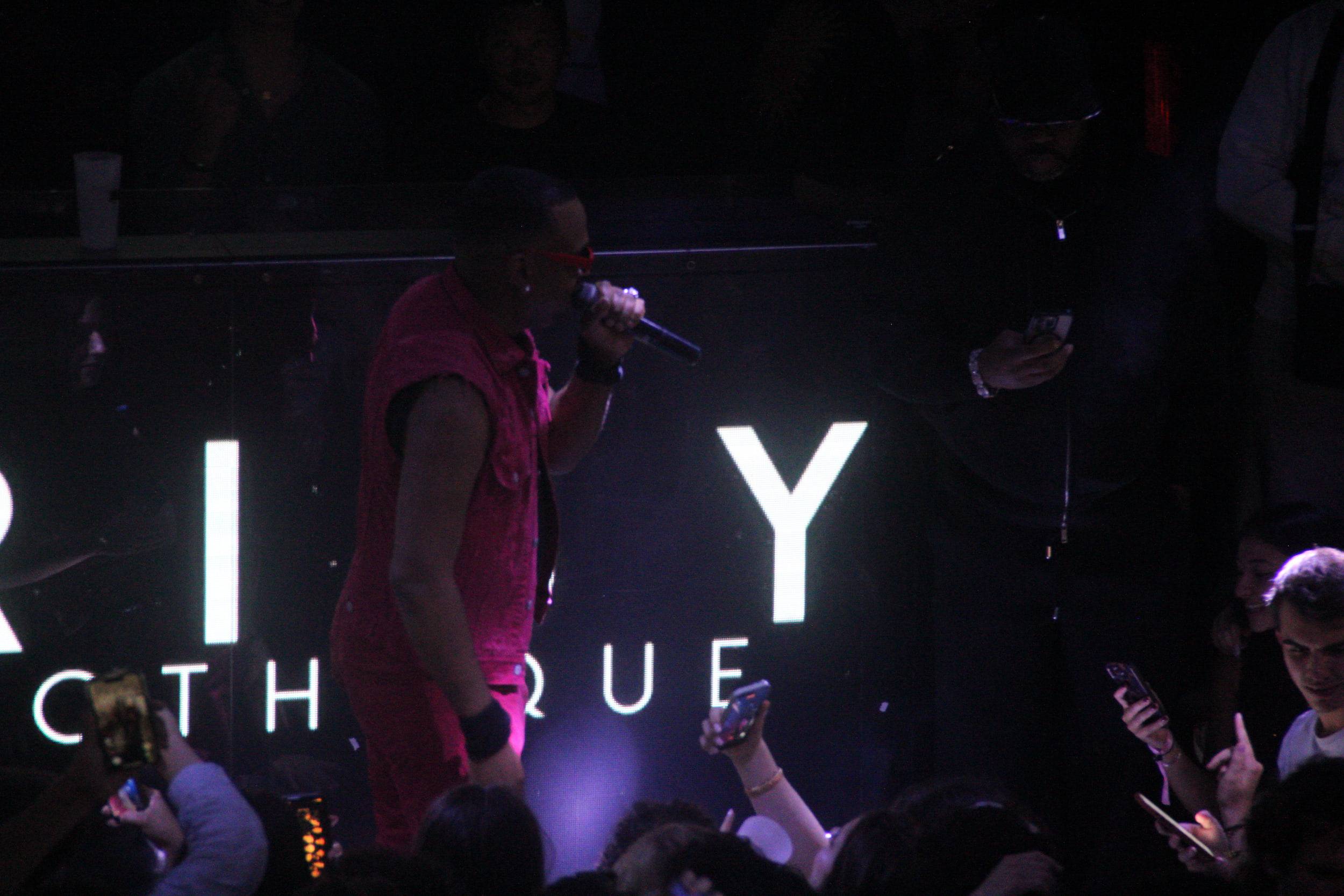 Un artiste en tenue rose chante au micro devant l'enseigne du club, des téléphones filment dans le public.