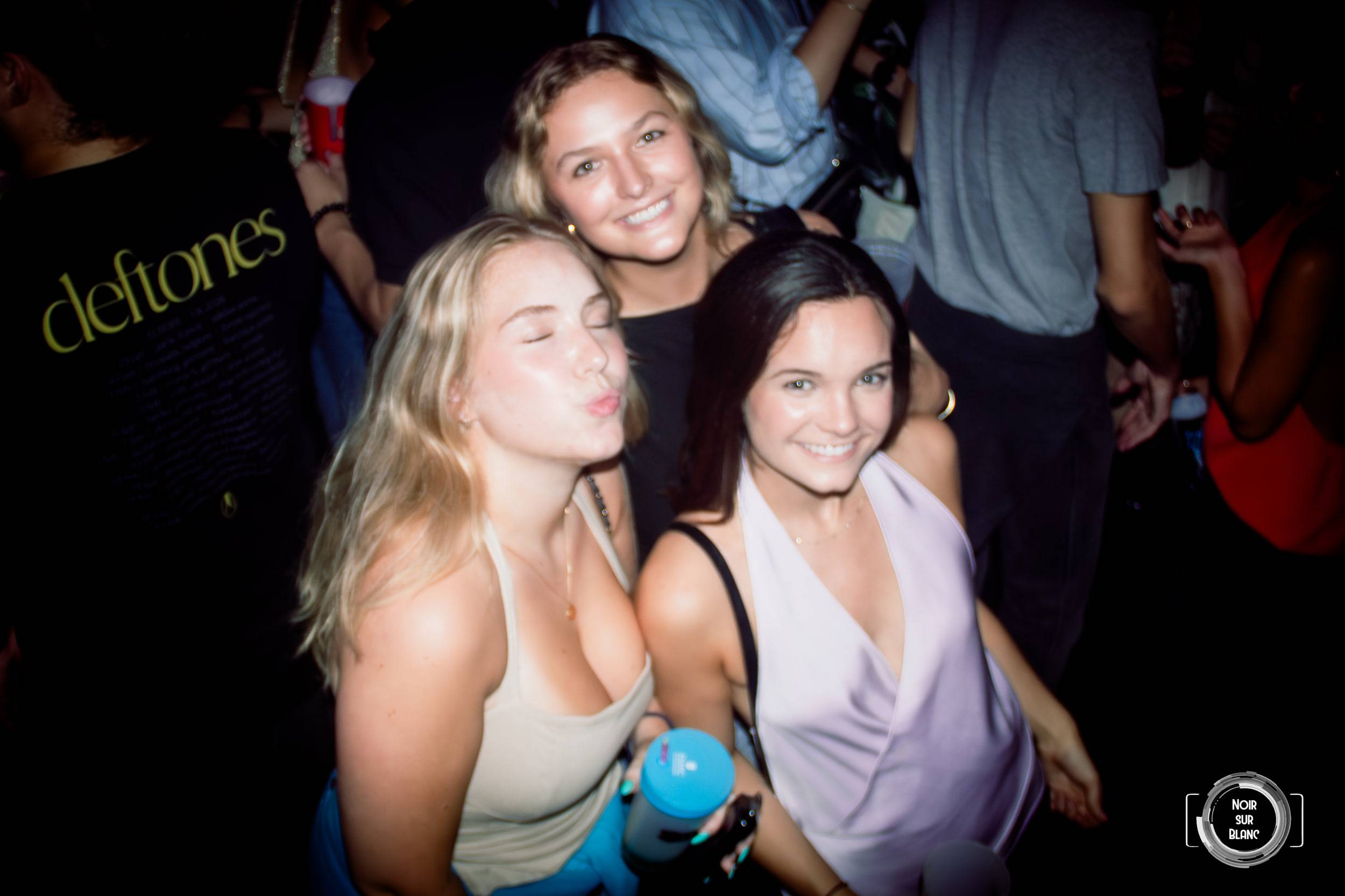 Trois jeunes femmes posent au flash, l'une envoyant un baiser à l'objectif, devant une affiche accrochée au mur.
