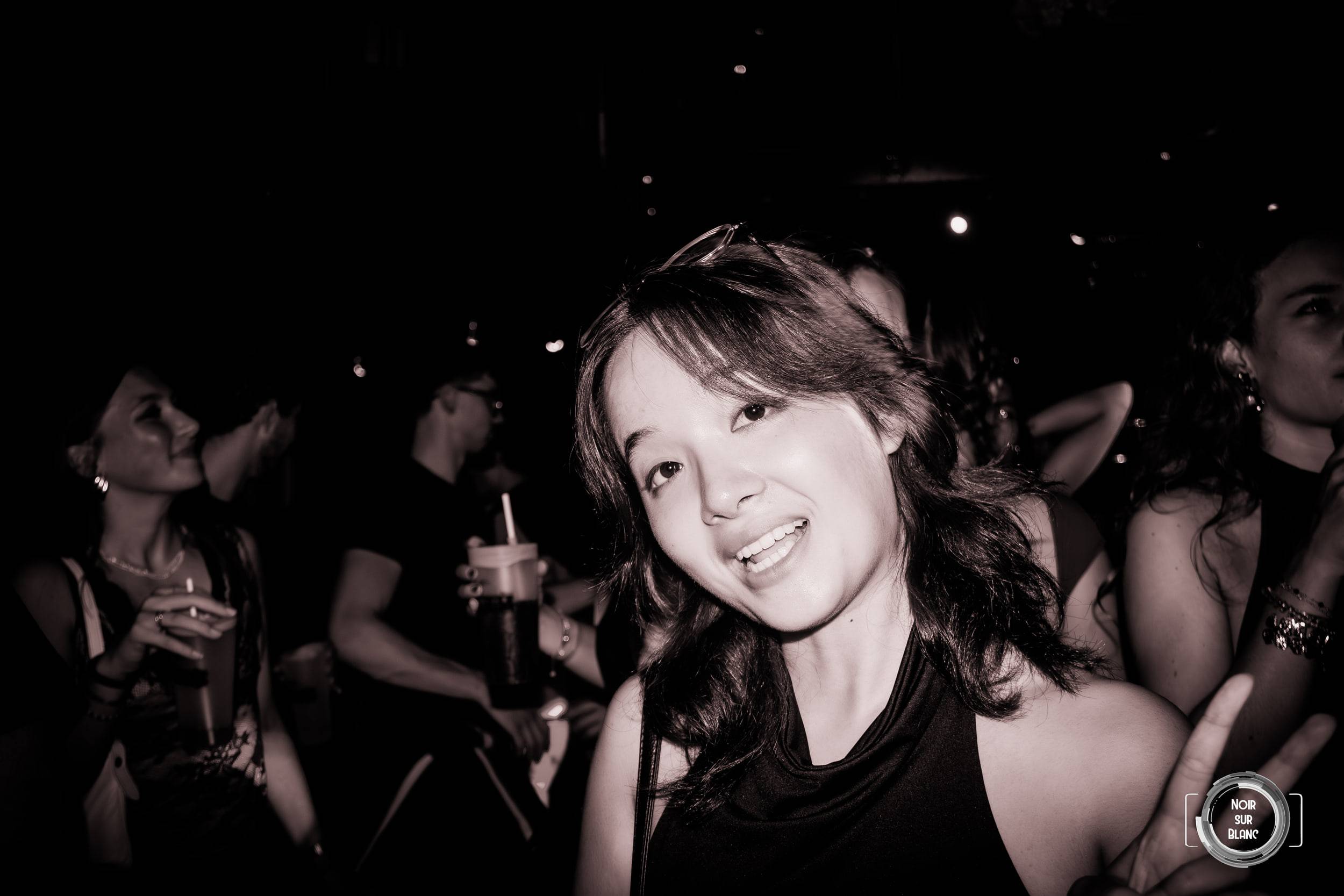 Photo en noir et blanc : une jeune femme sourit à l'objectif, saisie au flash au milieu d'une foule plongée dans le noir.