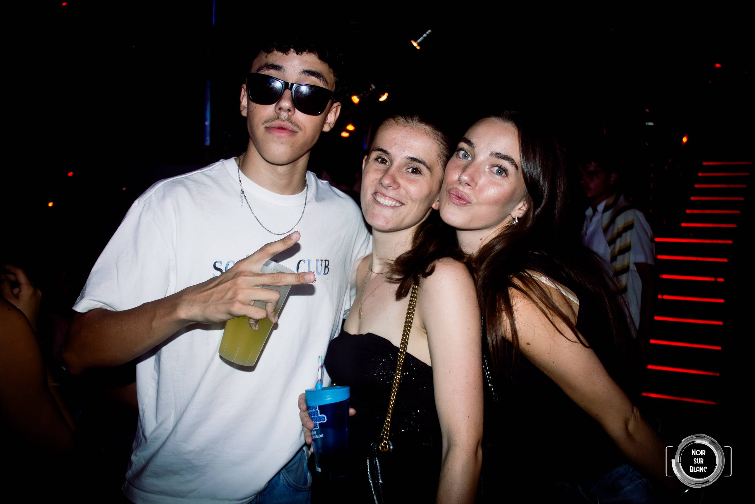Un jeune homme en lunettes de soleil pose entre deux jeunes femmes, un escalier bordé de LED rouges derrière eux.