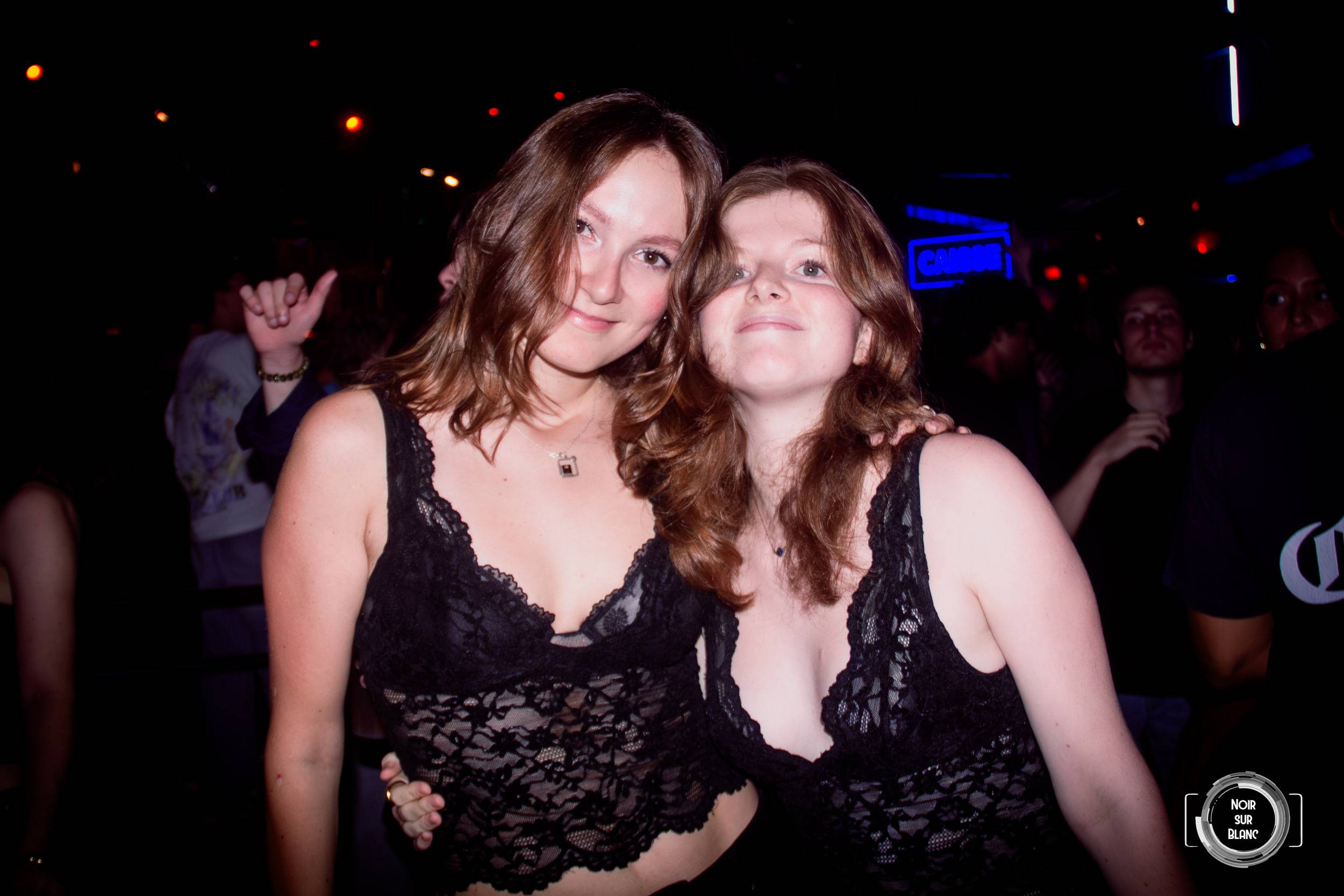 Deux jeunes femmes joue contre joue sourient à l'objectif, saisies au flash dans la pénombre du club.