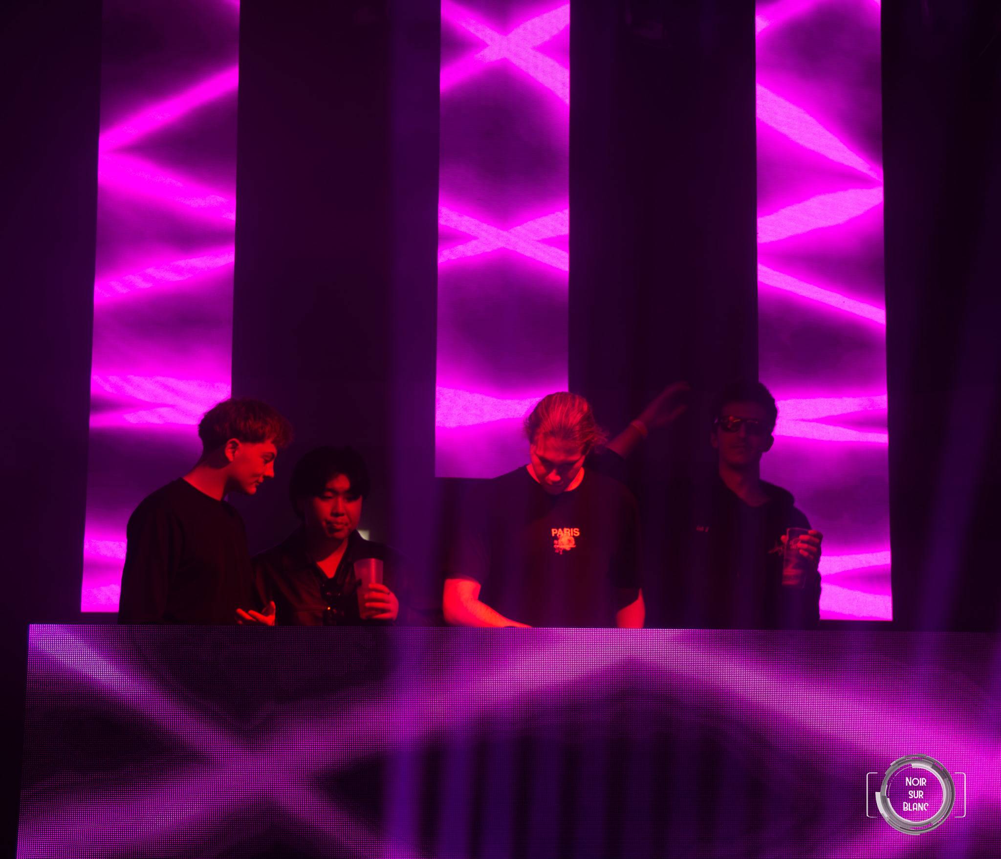 La scène baignée de néons roses en zigzag, quatre silhouettes derrière la console.