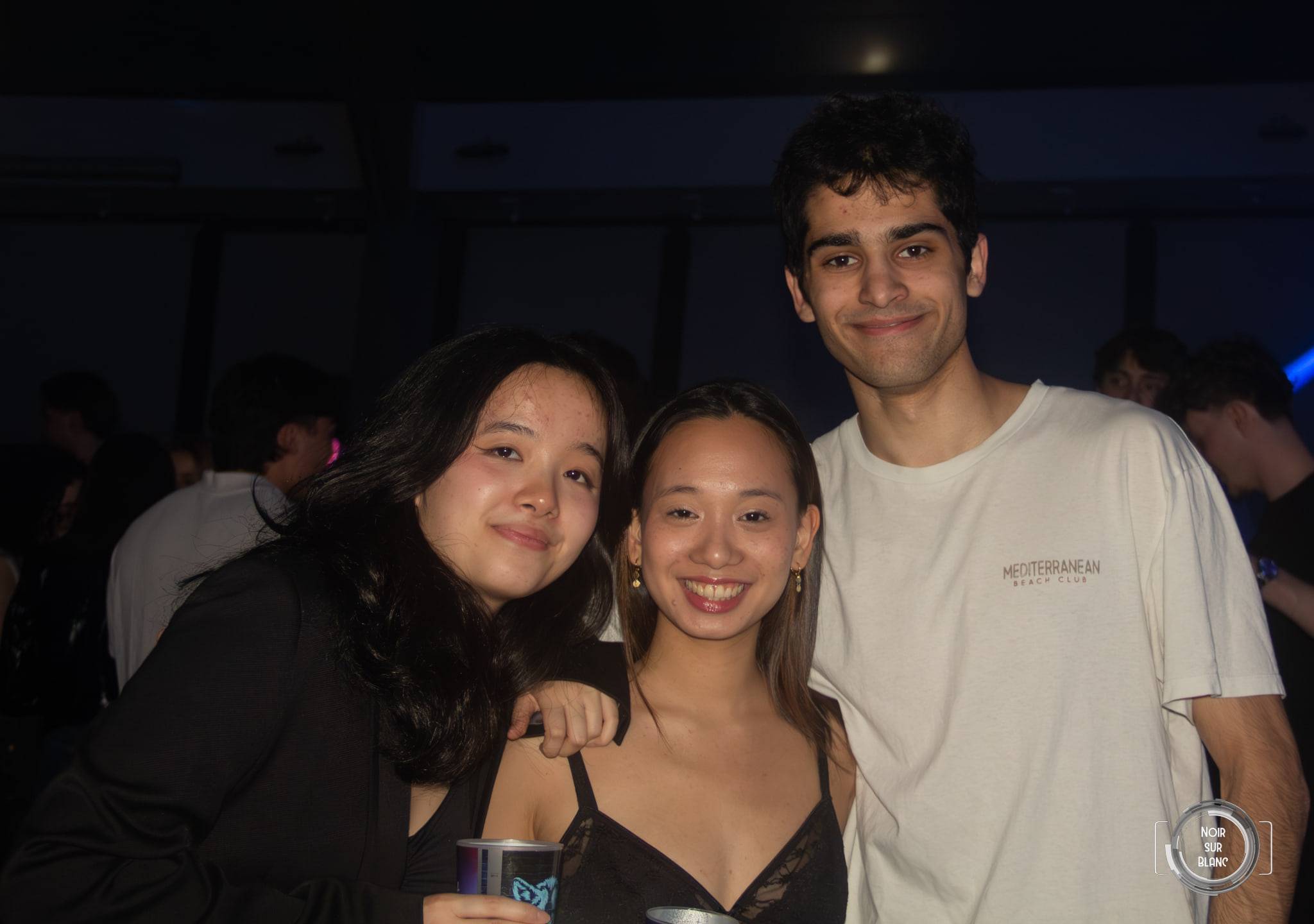 Trois amis posent souriants, gobelets à la main, dans la pénombre de la salle.