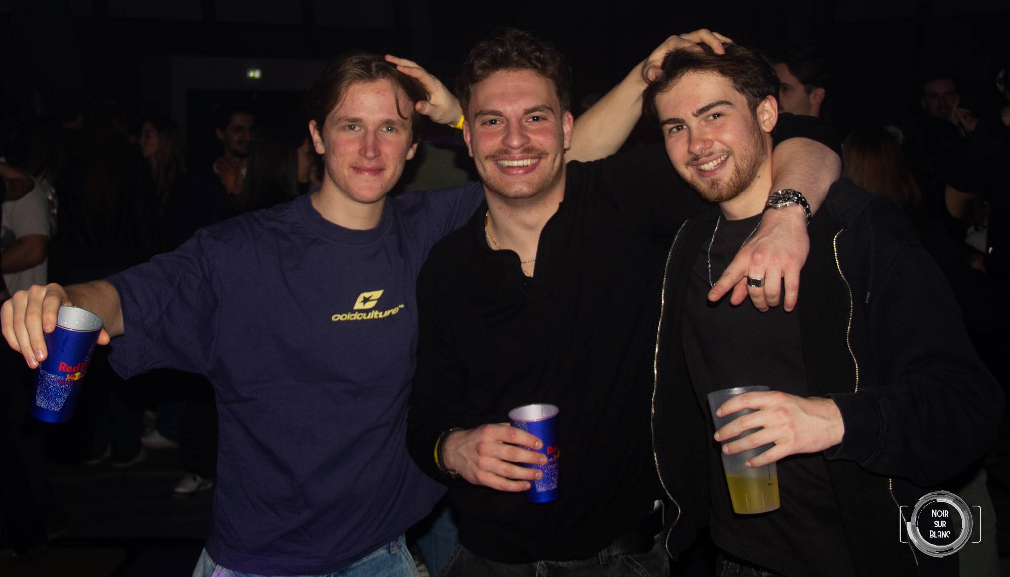 Trois étudiants se tapotent la tête en riant, gobelets à la main.