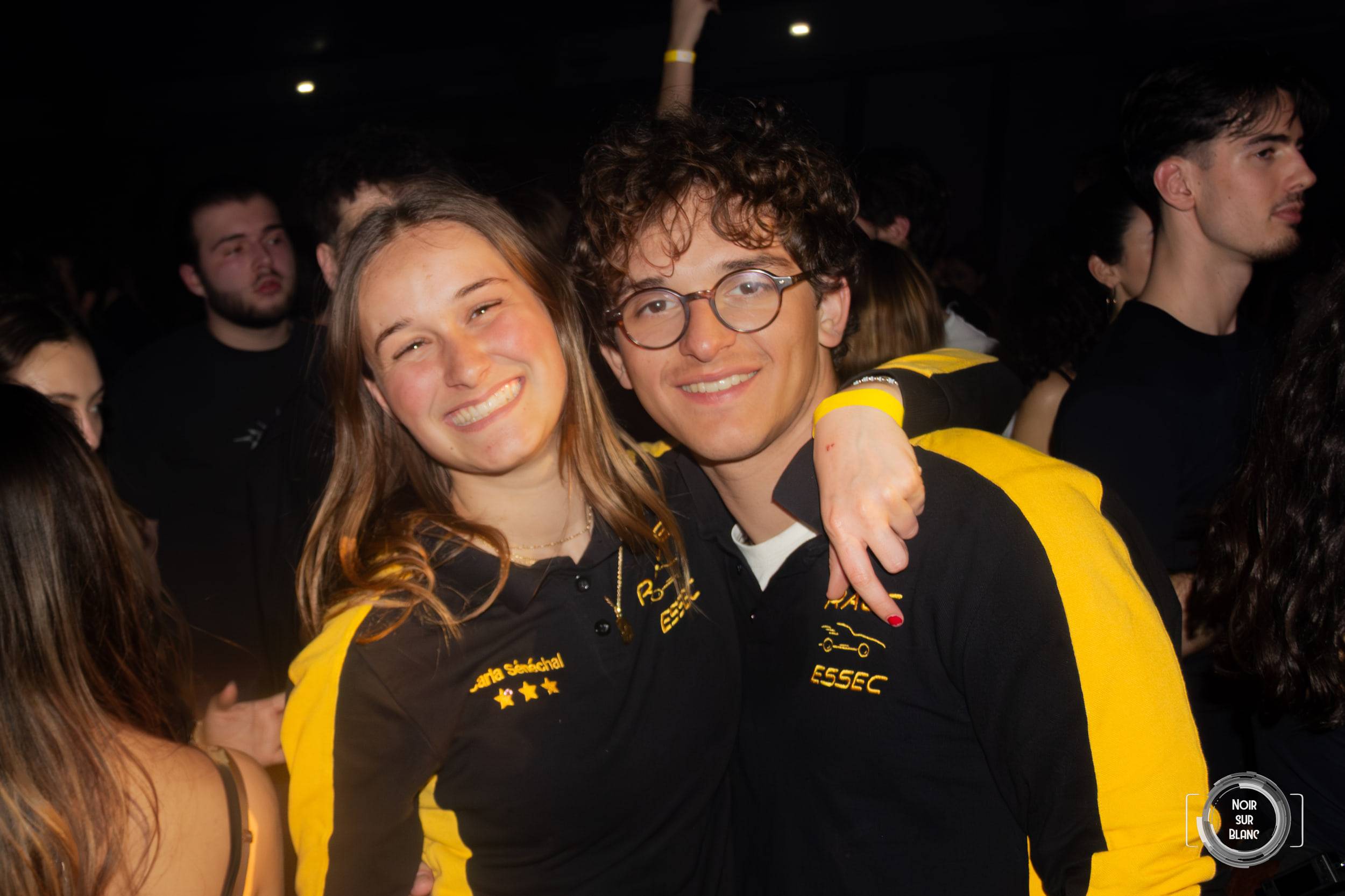 Une étudiante et un étudiant en veste jaune aux couleurs de l'ESSEC posent au milieu de la foule.