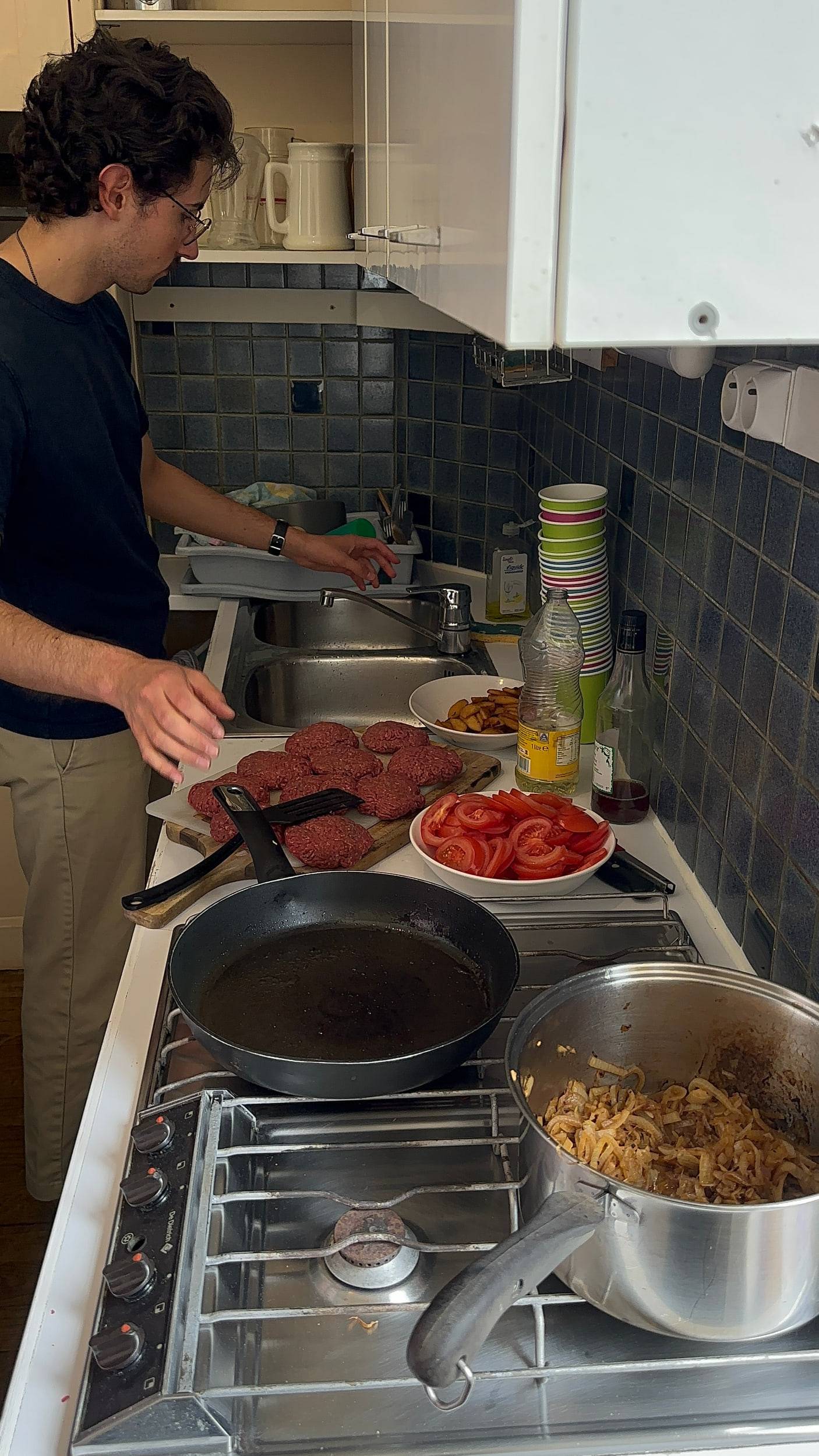 Quelqu'un prépare le repas dans une cuisine carrelée de bleu : planche de viande, tomates coupées, poêle et casserole sur le feu.