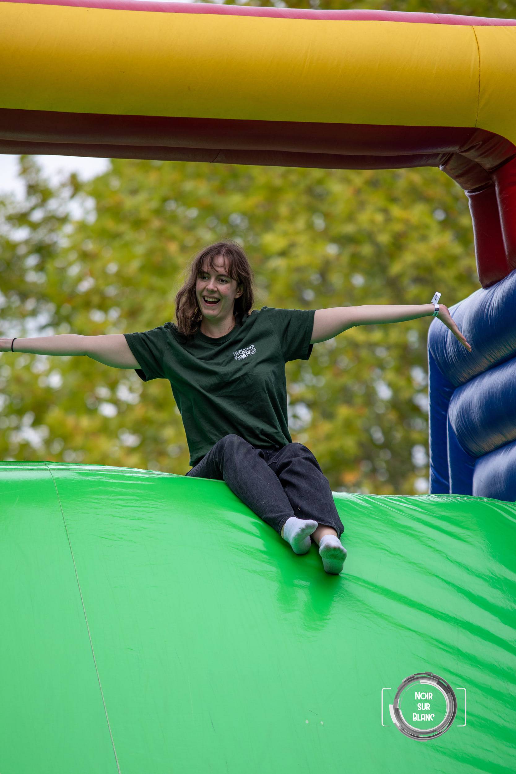 Une étudiante en t-shirt vert descend assise un toboggan gonflable, les bras écartés en croix, riant sous les arbres.