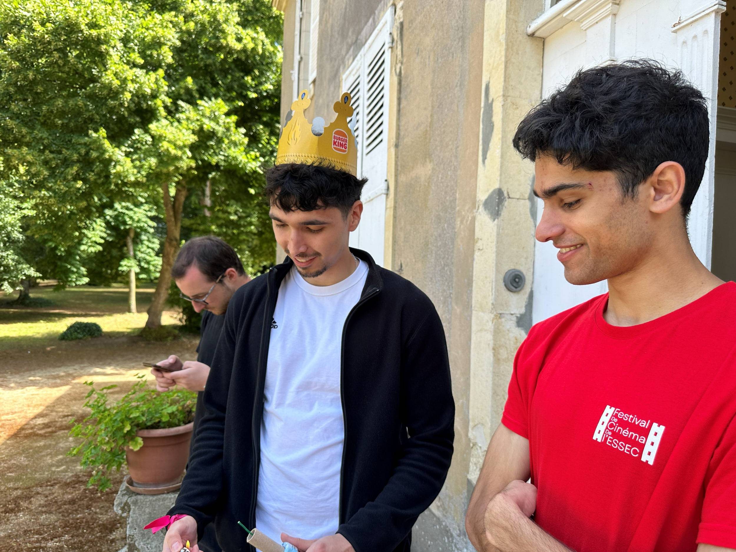 Deux membres discutent devant la façade de la maison, un troisième coiffé d'une couronne en papier les observe depuis le parc.