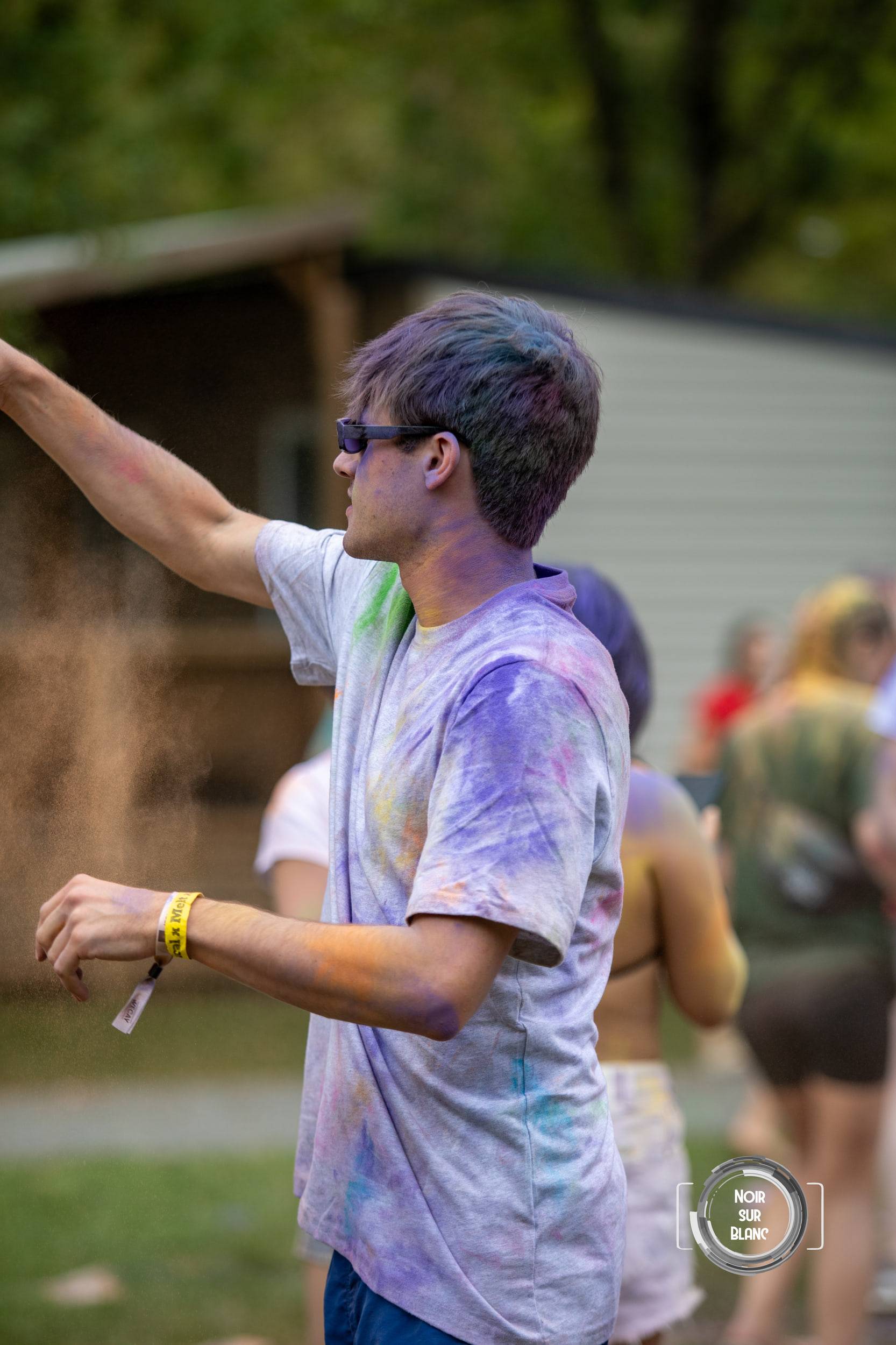 De profil, un étudiant aux cheveux et au t-shirt saturés de pigment violet lance une poignée de poudre, lunettes de soleil sur le nez.