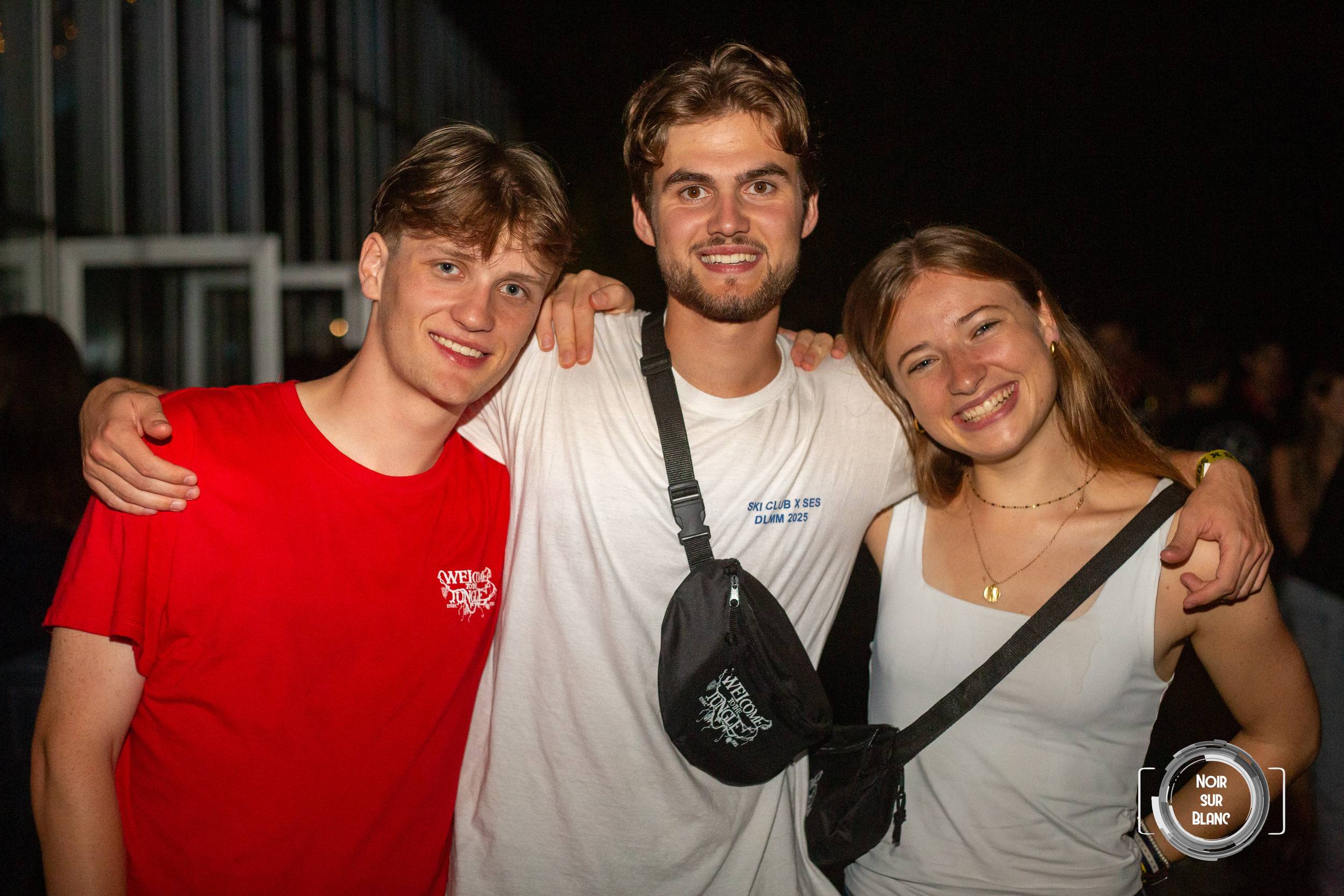 Trois étudiants posent bras sur les épaules dans la nuit, souriant face à l'objectif, une banane en bandoulière.