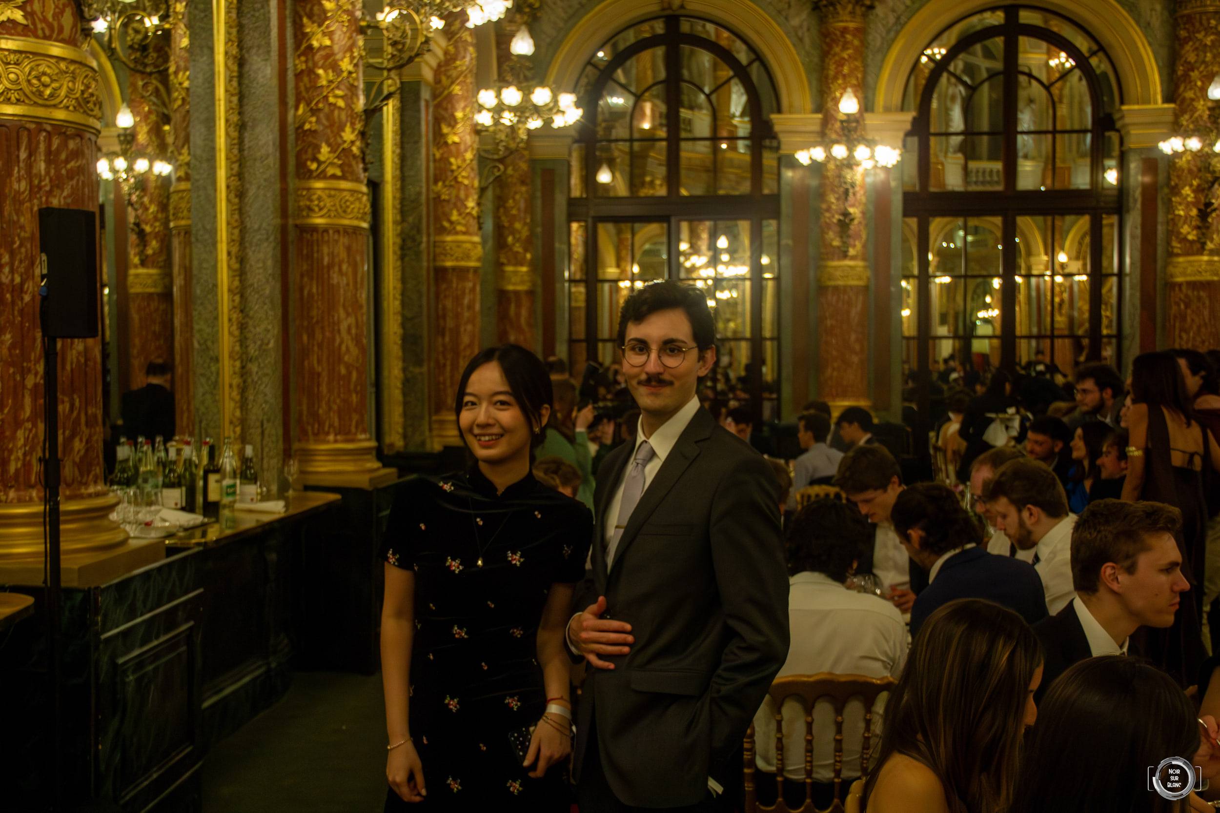 Un couple en tenue de soirée pose au milieu des colonnes dorées et des lustres d'une salle de bal bondée.