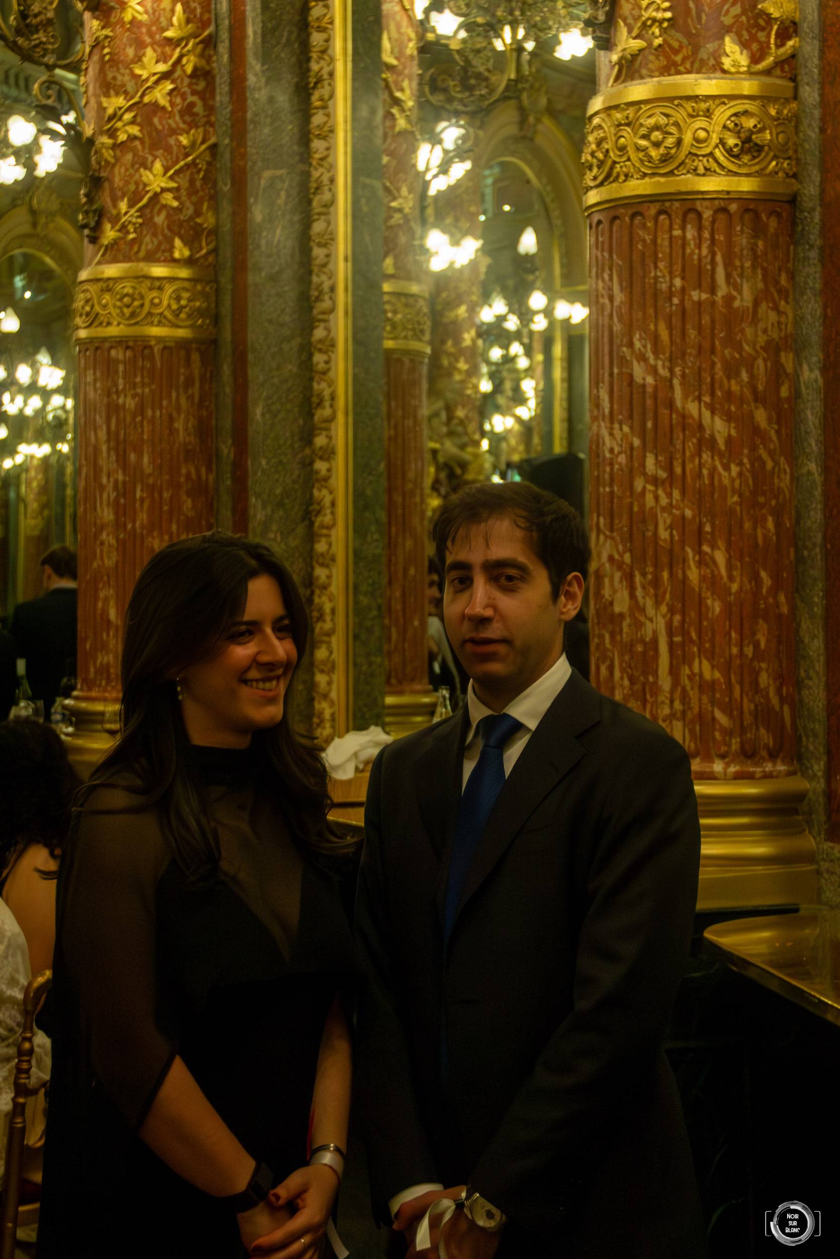 Une étudiante en noir et un étudiant en costume posent devant les marbres et les miroirs d'une salle de bal.
