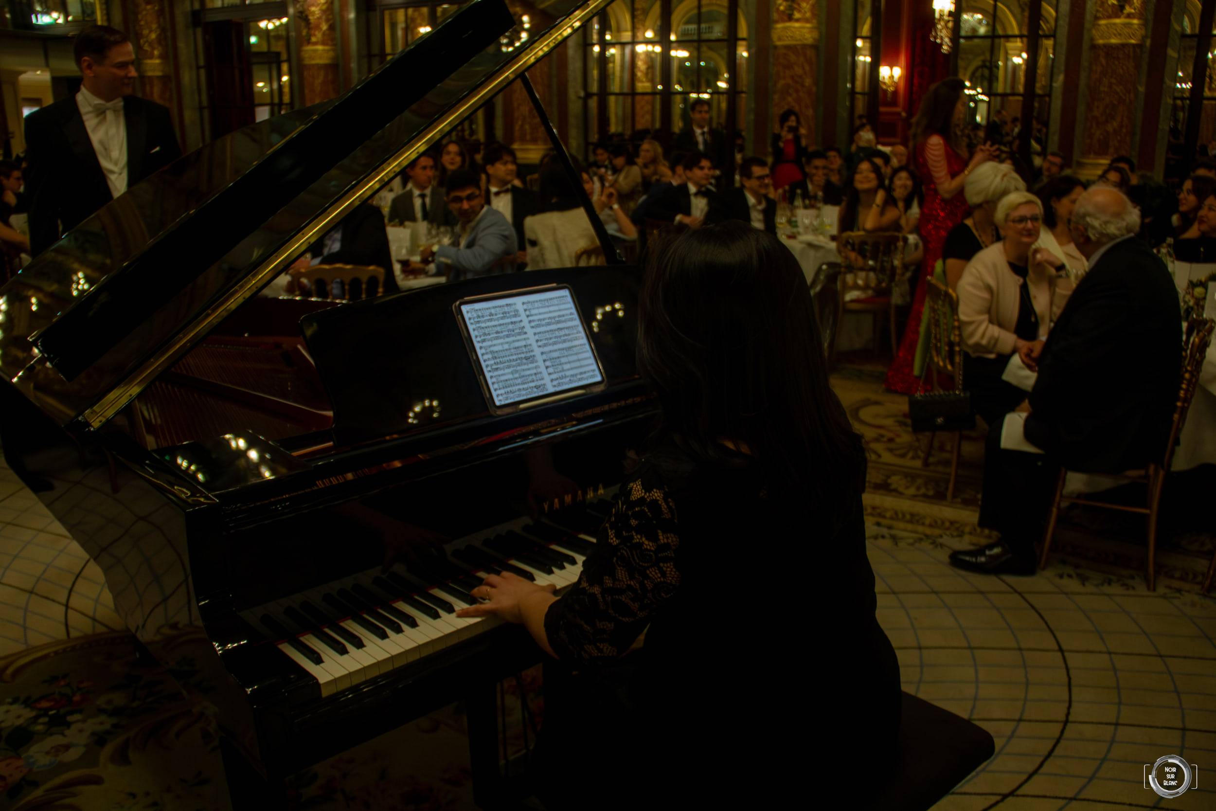 Un pianiste joue sur un quart de queue noir devant les tables du dîner, dans une salle aux plafonds peints.