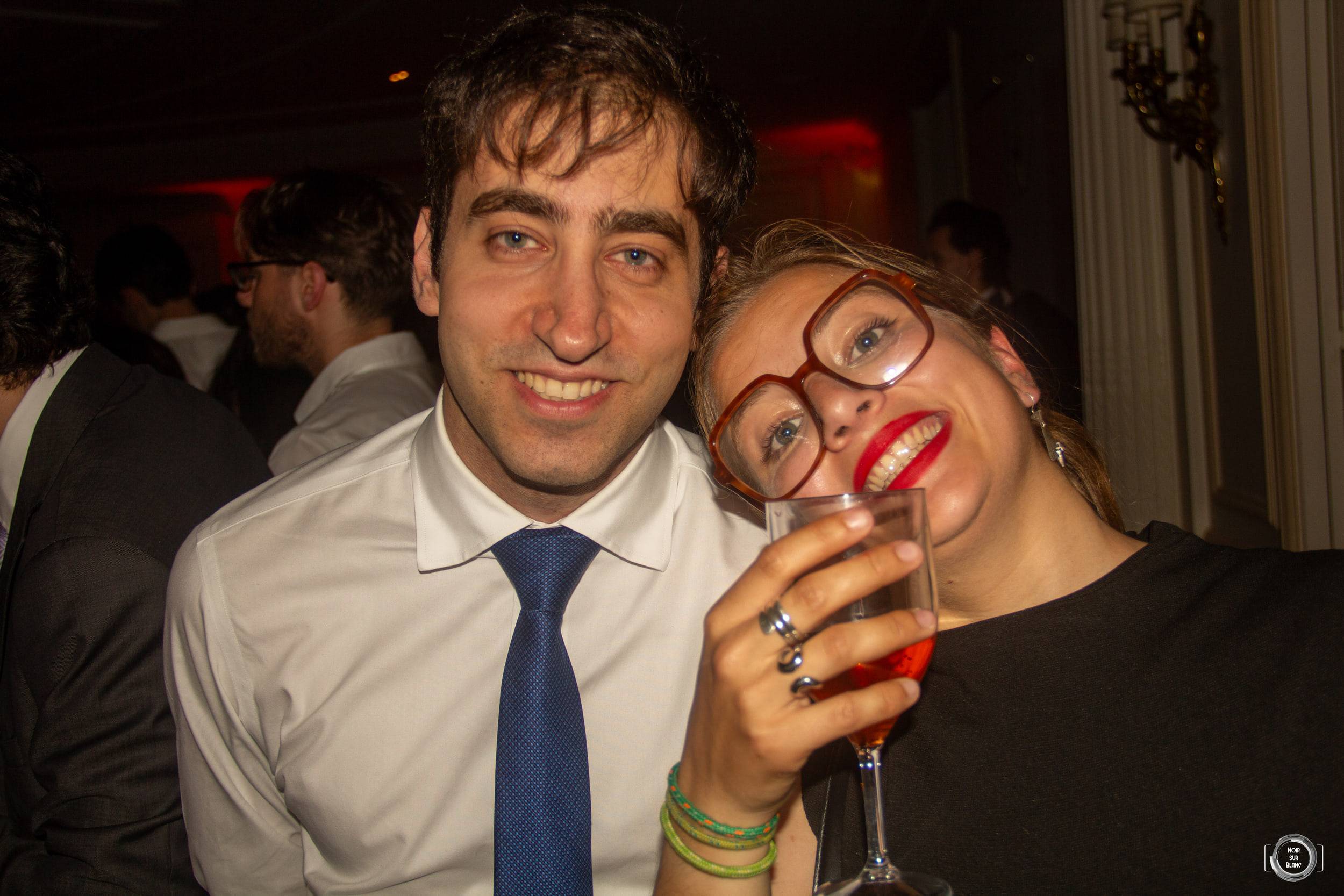 Un étudiant en chemise et cravate et une étudiante à lunettes posent joue contre joue, un verre levé devant l'objectif.