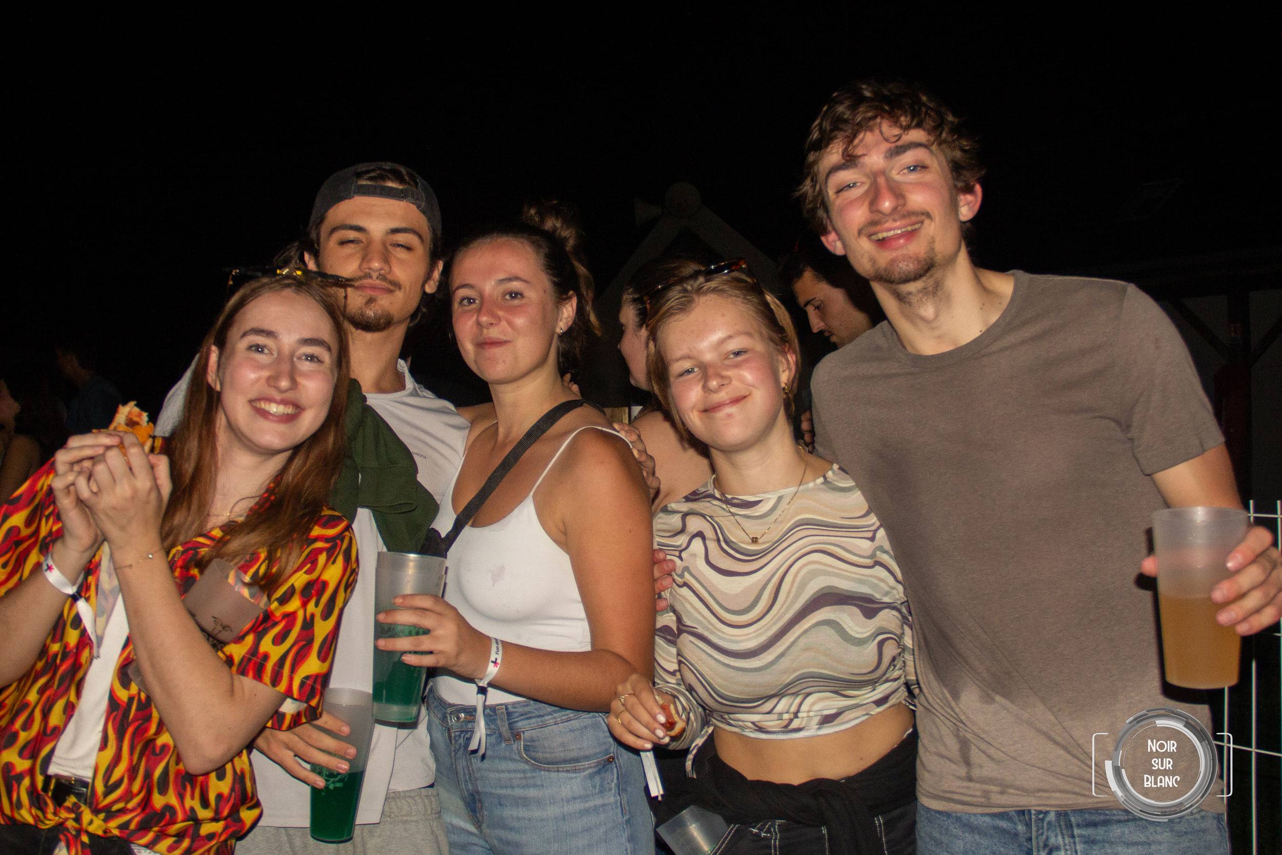 Cinq étudiants serrés les uns contre les autres posent dans la nuit, gobelets à la main, l'une portant une chemise à motif de flammes.