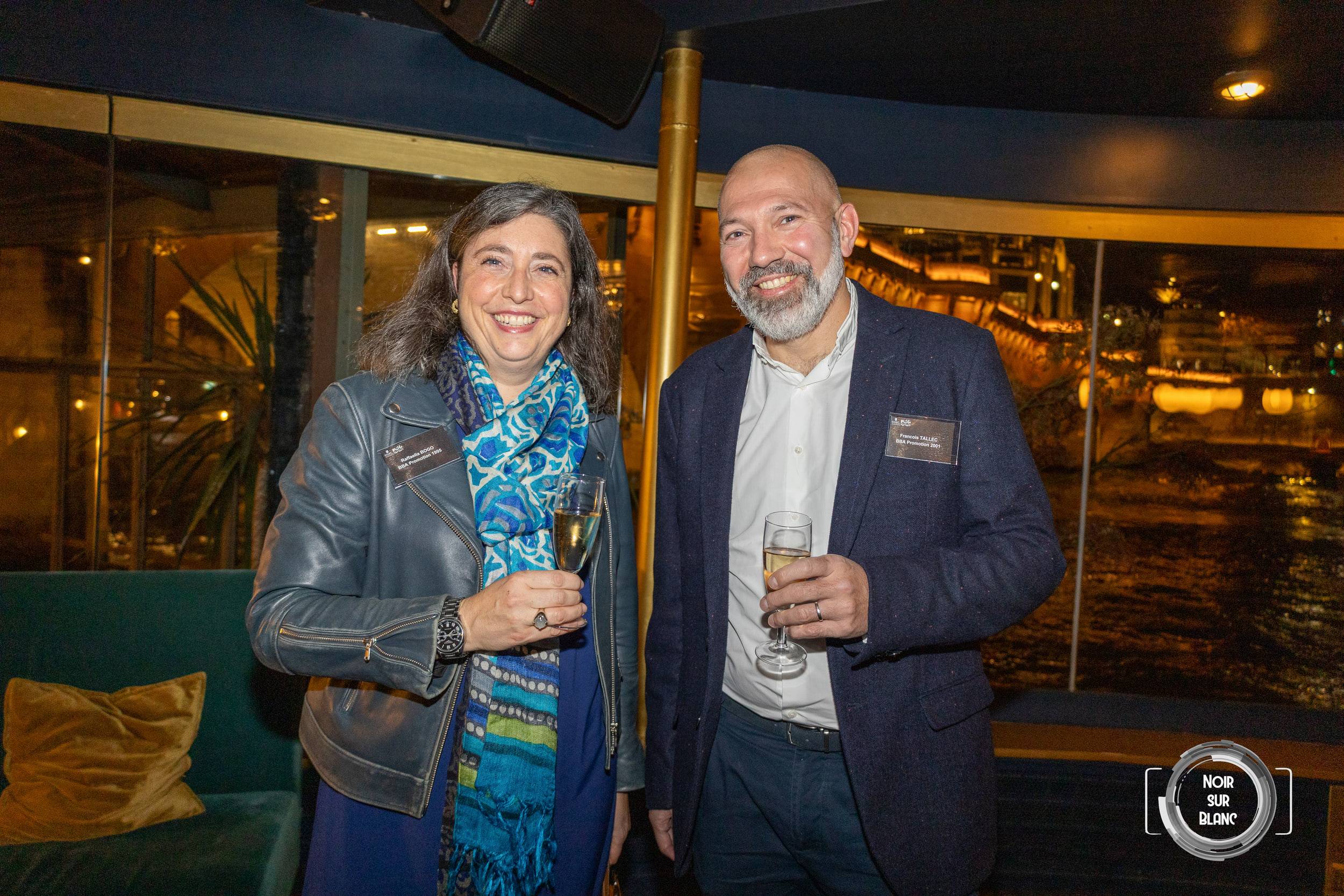 Une invitée et un invité en veste posent côte à côte, coupe de champagne à la main, à bord d'une péniche.