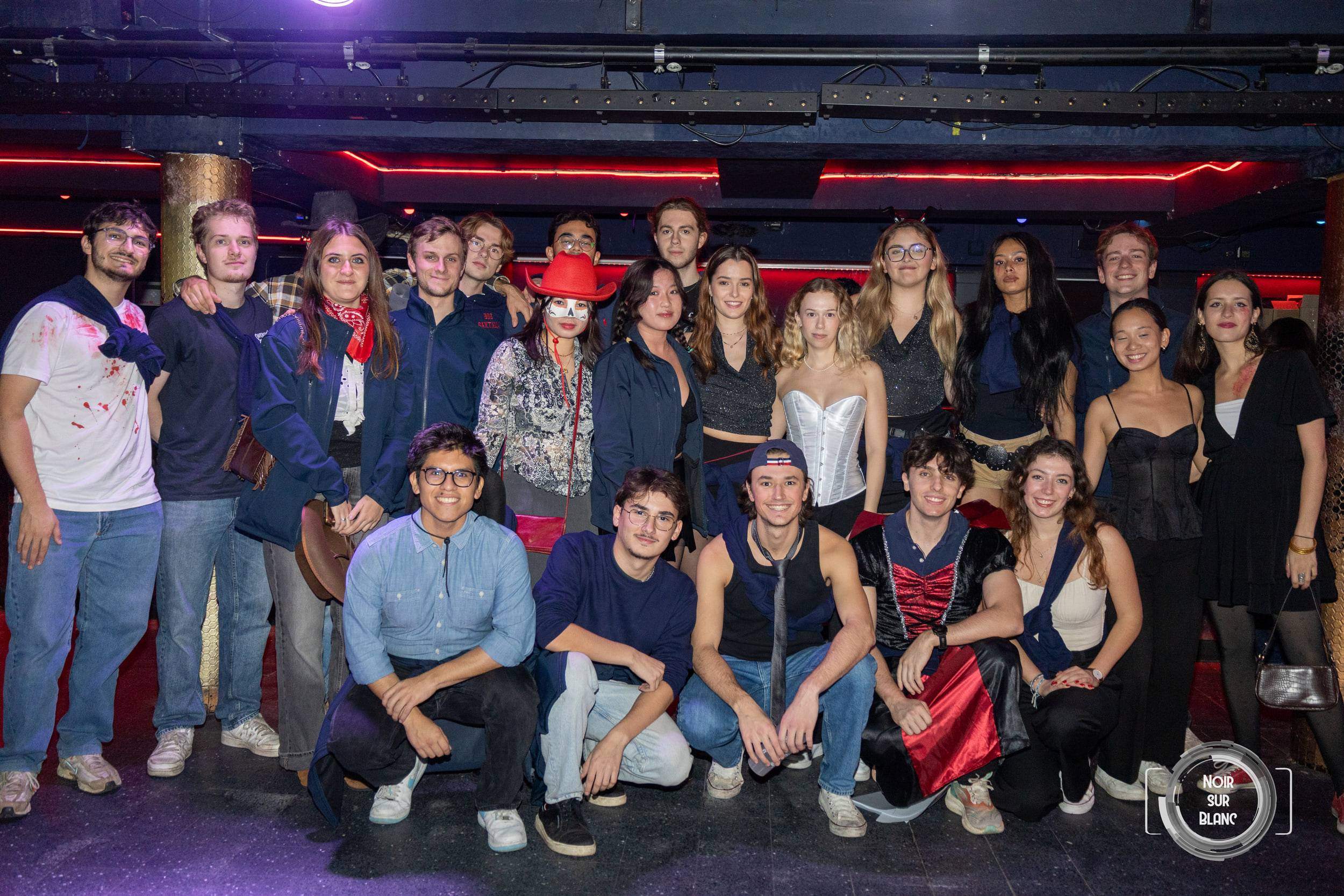 Photo de groupe en deux rangées, debout et accroupis, une partie des étudiants encore déguisés, devant les néons rouges de la salle.