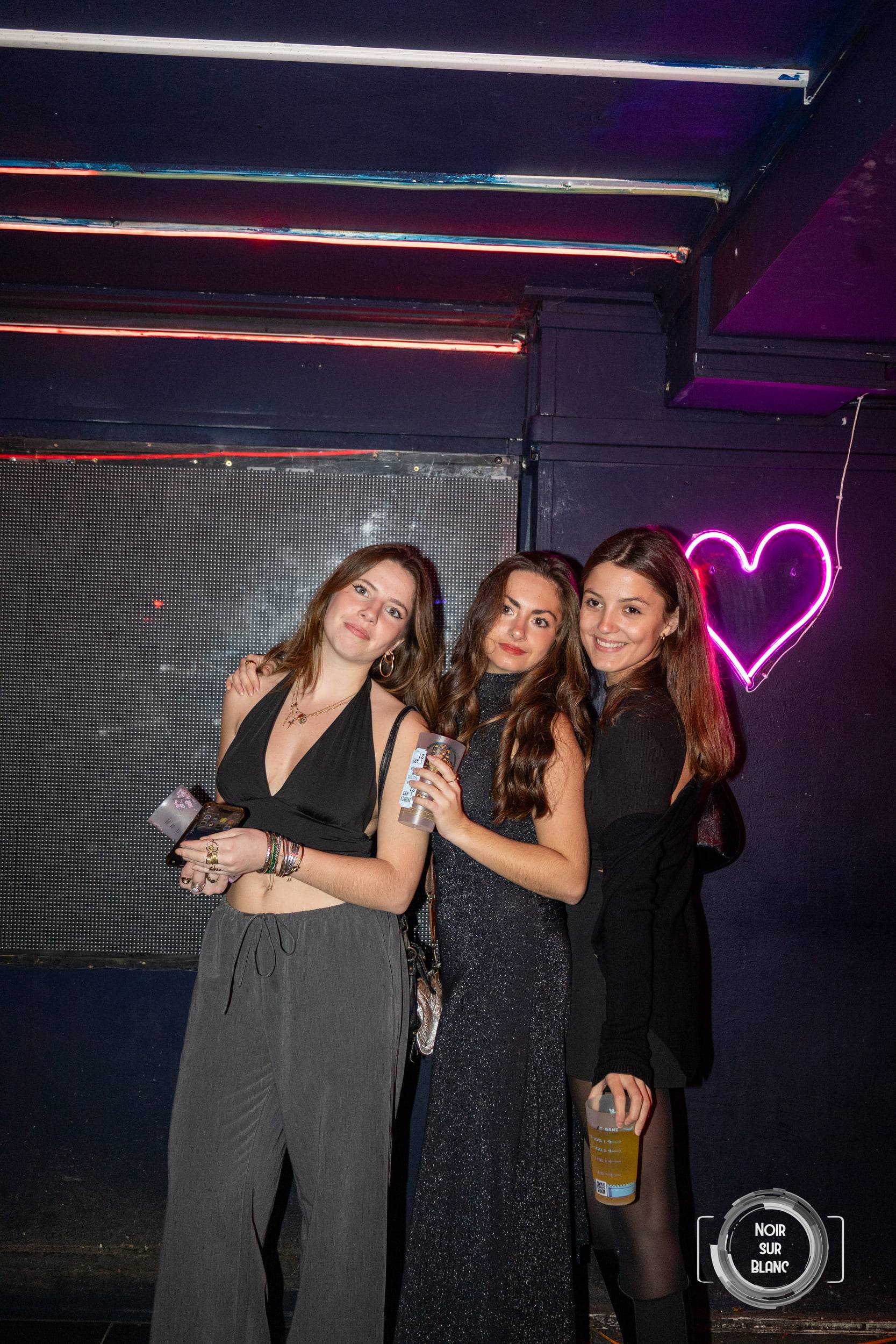 Trois amies posent enlacées devant un cœur en néon rose accroché au mur sombre du club.