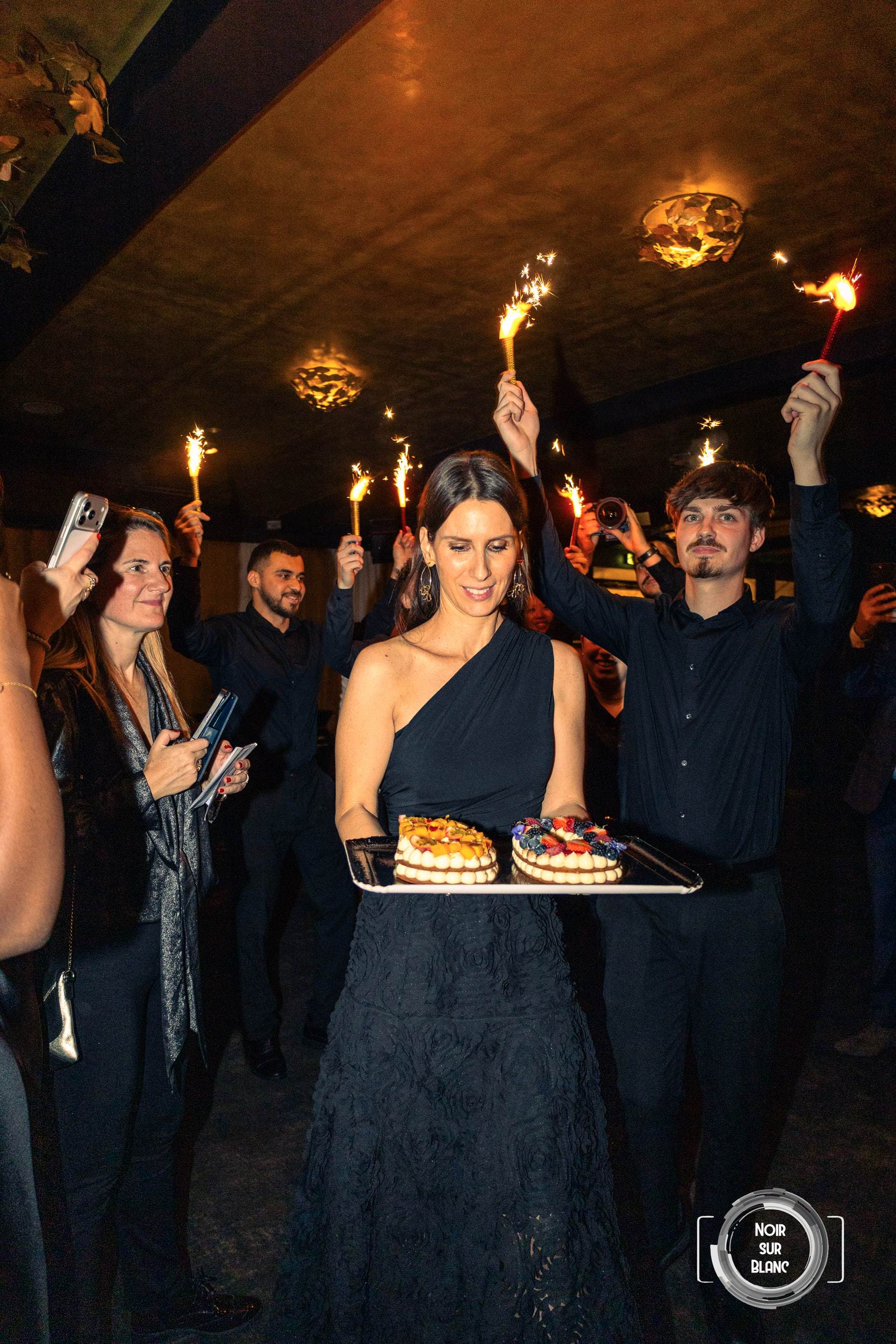 Au milieu d'une haie de cierges magiques brandis à bout de bras, une femme en robe noire porte deux gâteaux d'anniversaire sur un plateau.