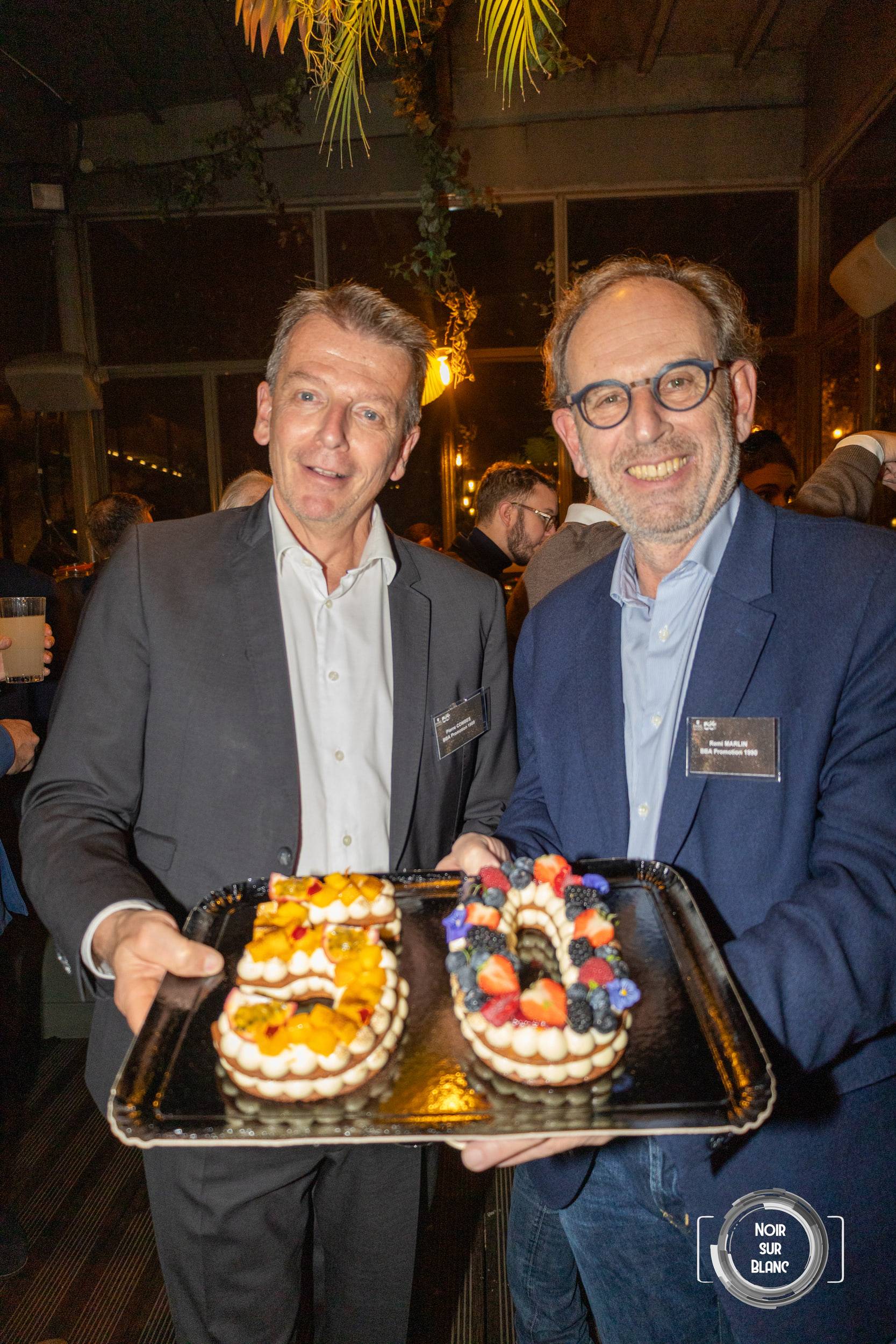 Deux hommes en costume présentent à l'objectif un plateau portant deux gâteaux en forme de 5 et de 0.