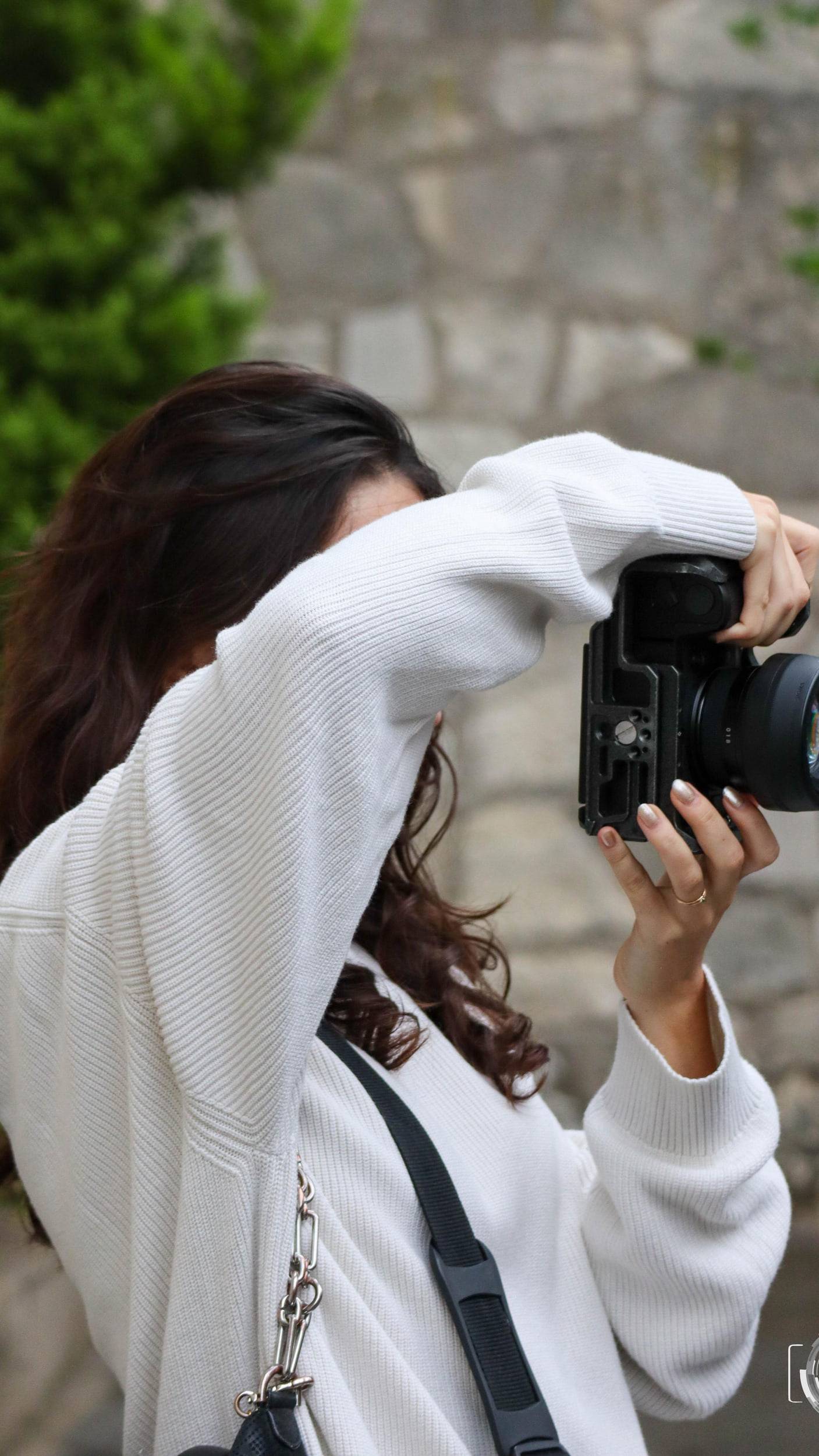 Une photographe de dos, en pull blanc, cadre une scène avec son appareil devant un mur de pierre.