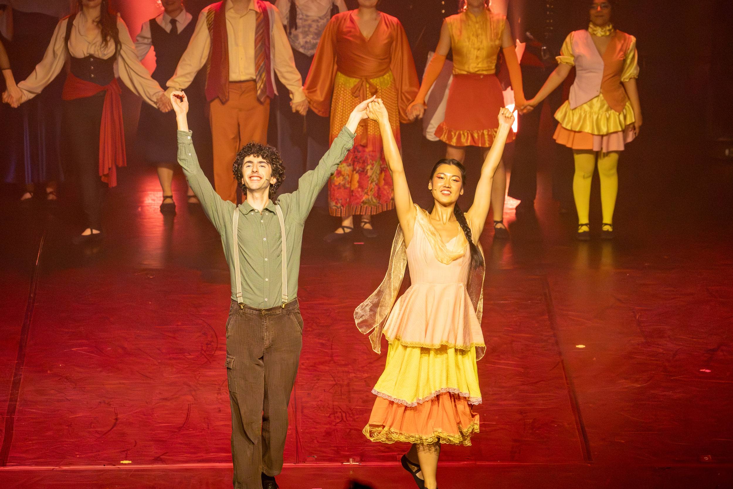 Deux comédiens saluent bras levés, la troupe alignée derrière eux dans la lumière rouge.