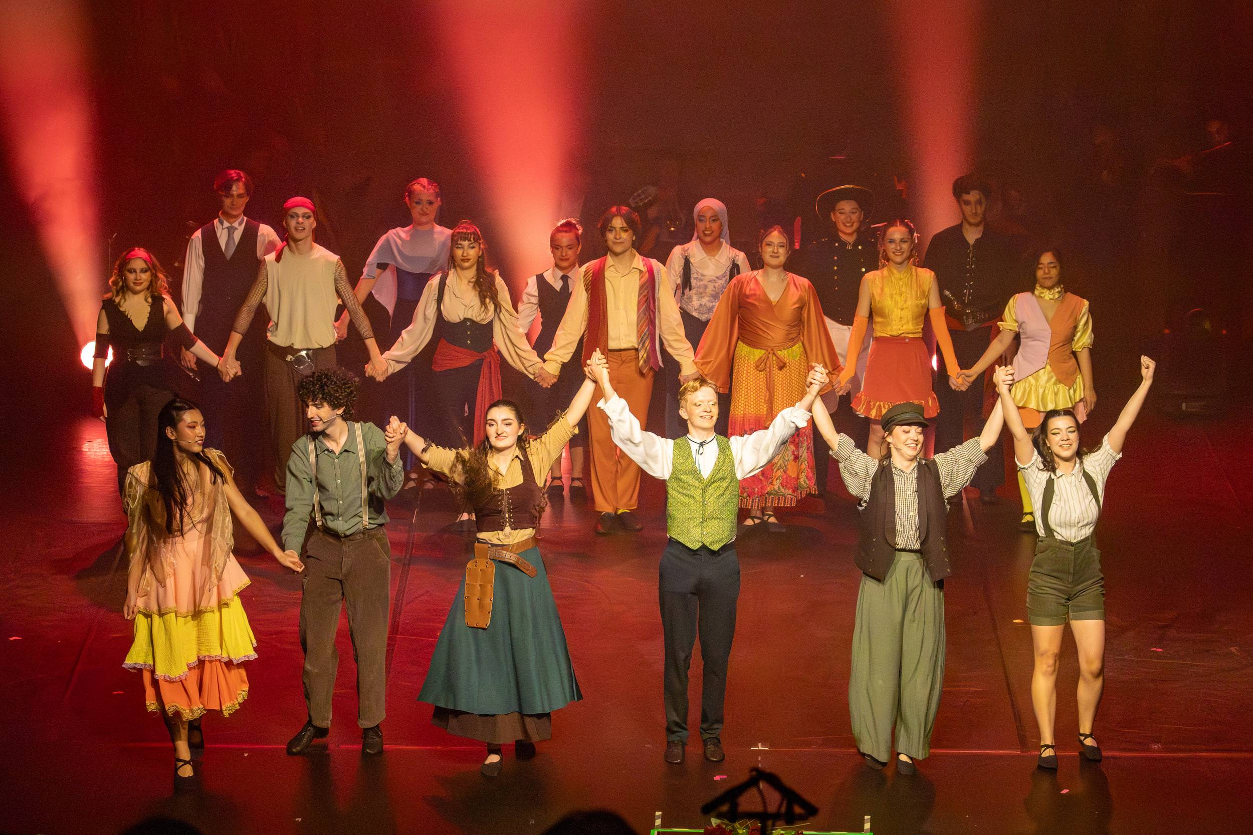 La troupe se tient par la main pour saluer, sur deux rangs, dans une lumière rouge.