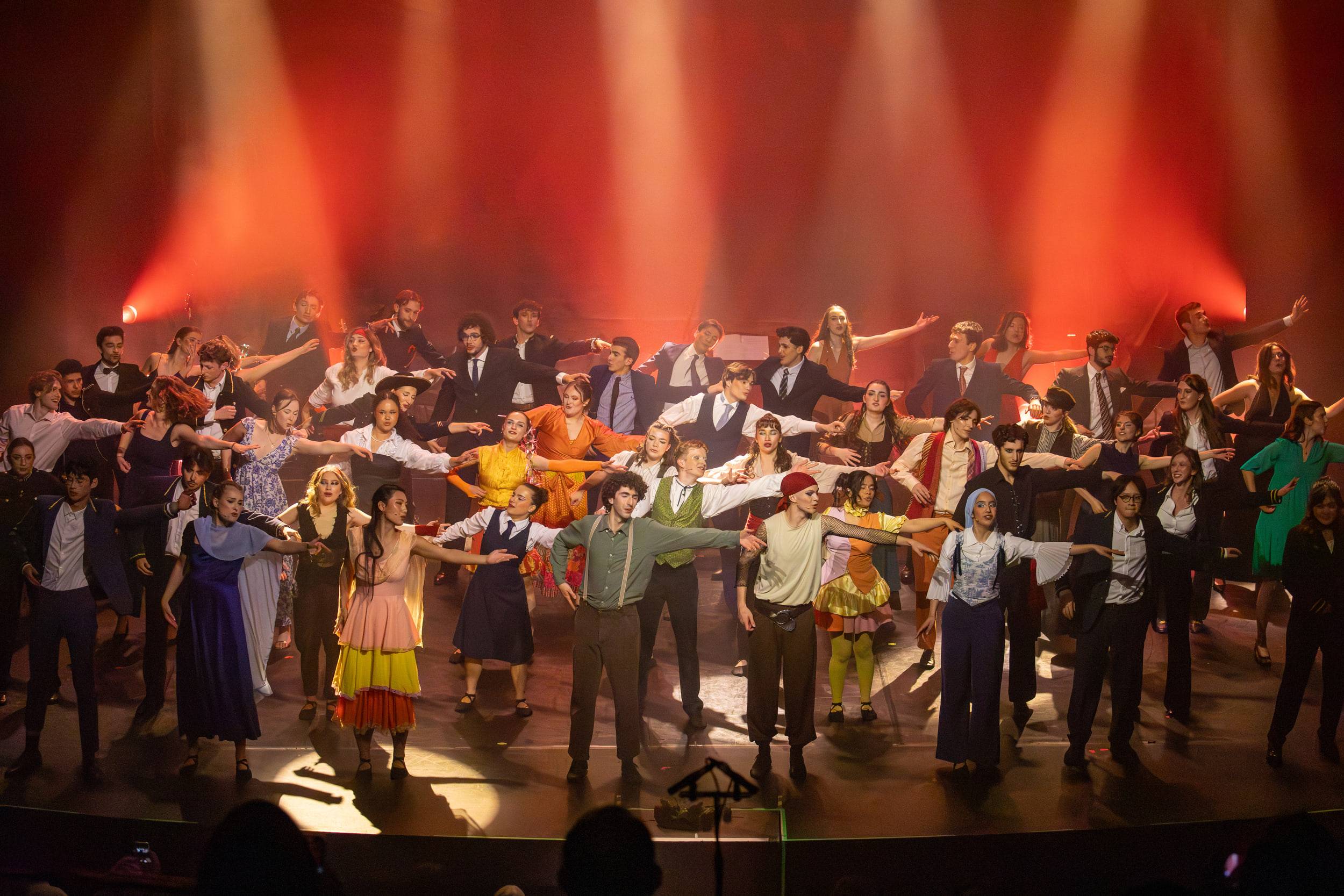 La troupe au complet, bras écartés, occupe toute la largeur du plateau sous une lumière rouge.