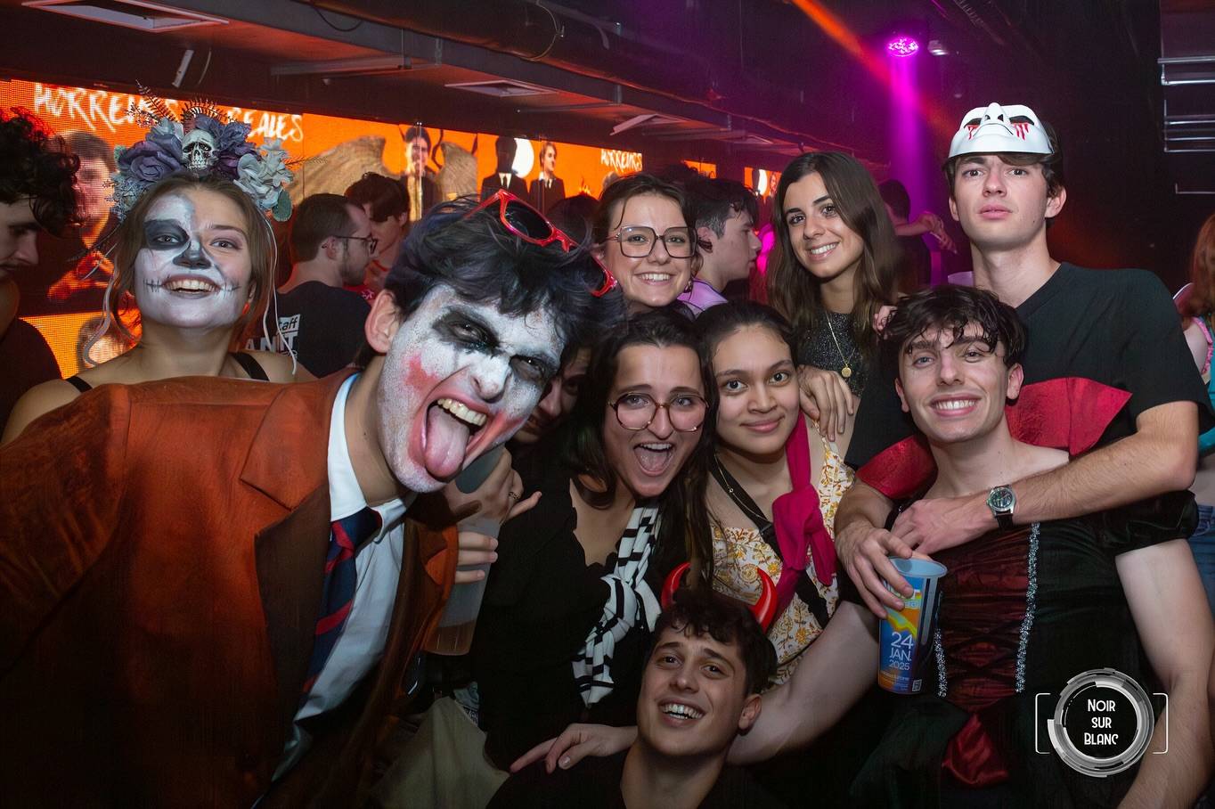 Un groupe d'étudiants déguisés et maquillés pour une soirée Halloween, serrés devant l'objectif dans une salle éclairée en orange.
